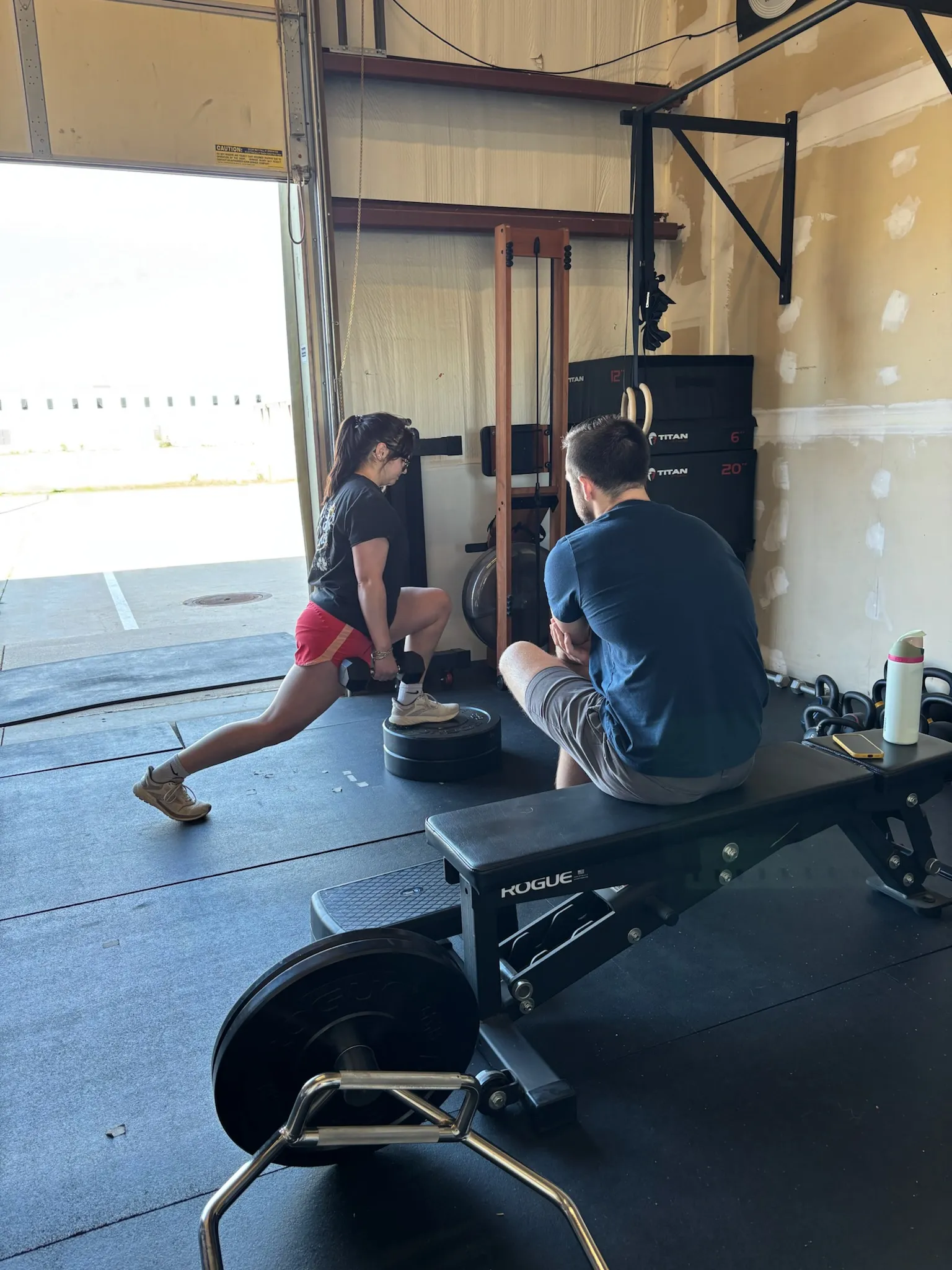 Woman performing a weighted step-up exercise in a gym while a man sits on a bench watching her.