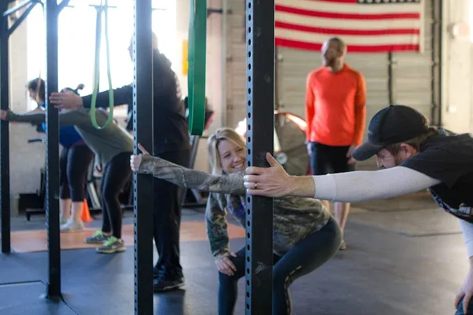 A group of people stretching inside a gym, holding onto metal bars while bending their upper bodies forward.