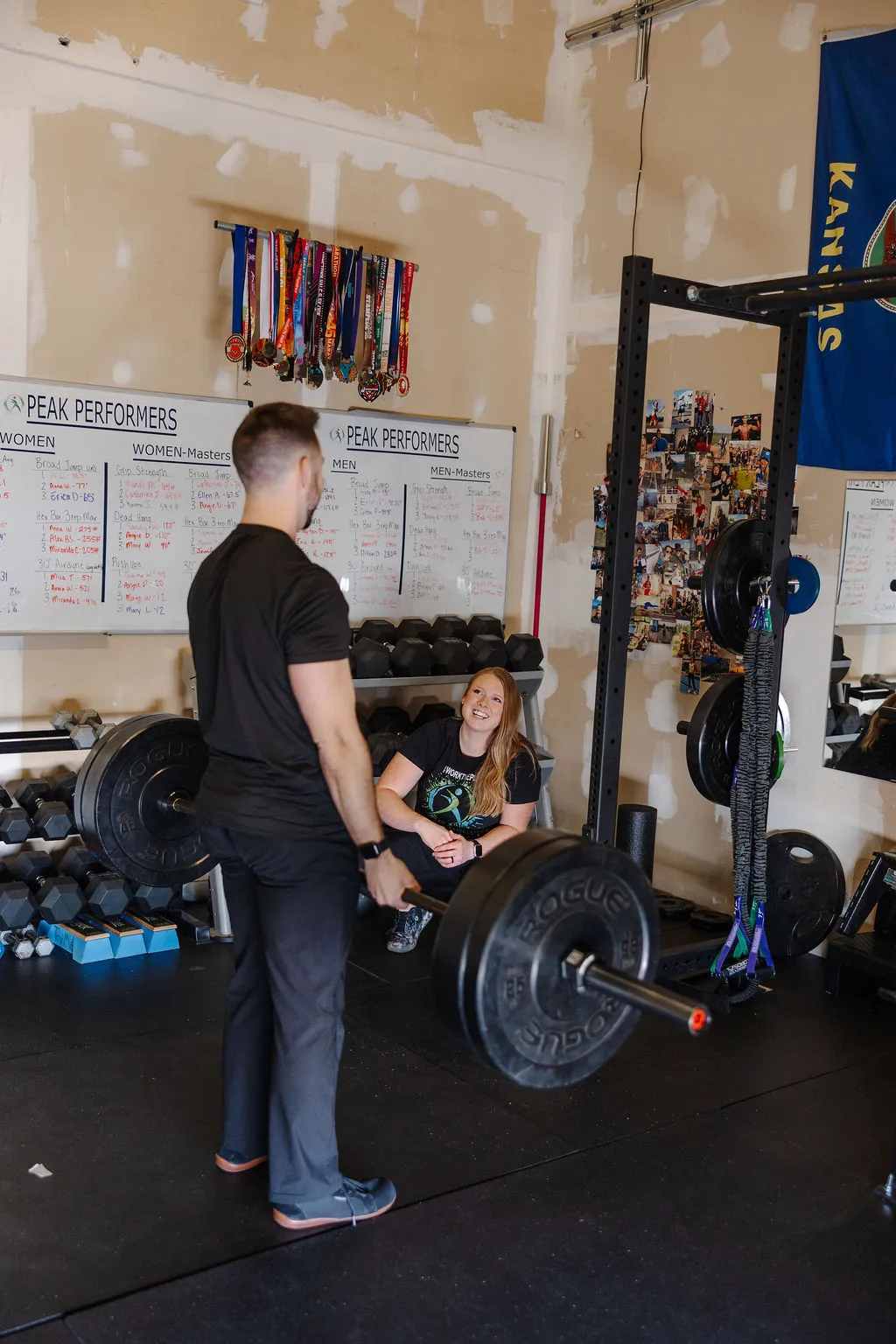 Man lifting a heavy barbell in a gym while a woman, sitting on the floor, watches and smiles.