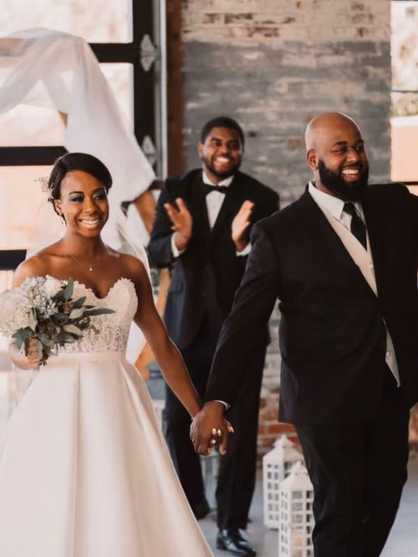 Man in suit pops champagne bottle while woman in white dress hugs him happily outdoors.