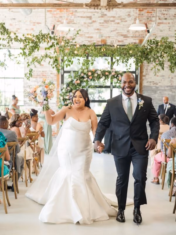 Man in suit pops champagne bottle while woman in white dress hugs him happily outdoors.
