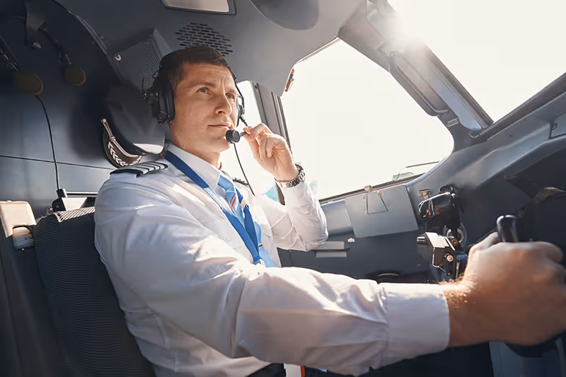 Male pilot in uniform wearing a headset, flying a private plane in the cockpit during daylight.
