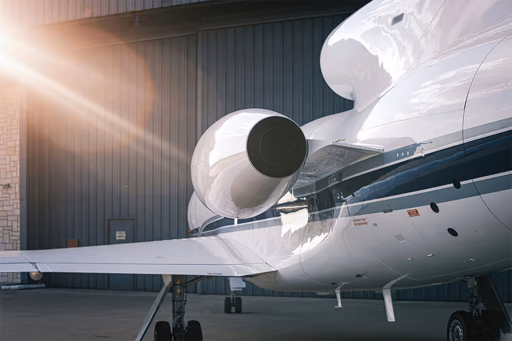 Close-up of a private jet engine and wing parked inside an aircraft hangar with sunlight shining from the top left.