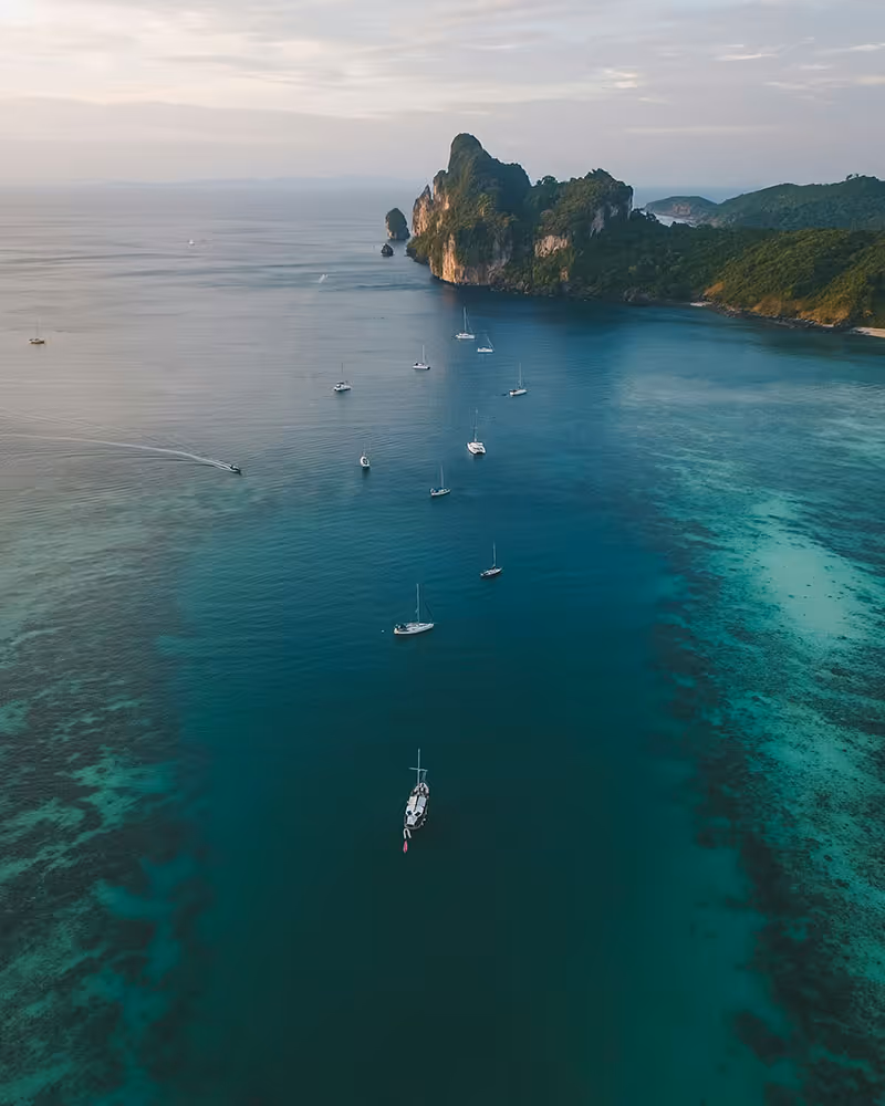 Aerial view of sailboats anchored in clear blue water near a lush rocky island at sunset.