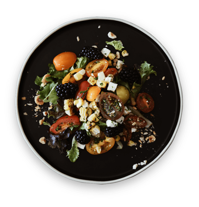 Mixed green salad with cherry tomatoes, blackberries, grilled corn, feta cheese, and chopped nuts on a black plate.