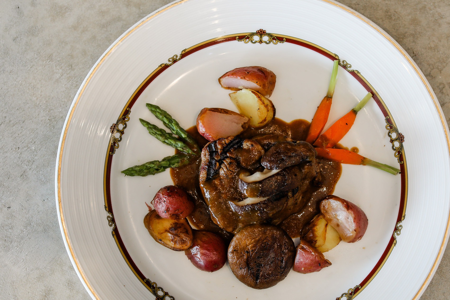 Plate with grilled steak topped with sautéed mushrooms, served with baby carrots, asparagus spears, and roasted red potatoes on a decorative white plate.