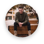 Young man wearing a dark green sweater, black pants, and light brown boots sitting on a wooden box outdoors with blurred building and lights in the background.
