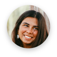 Smiling young woman with long dark hair wearing earrings and a light-colored top, photographed in natural light near a textured wooden surface.