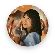 Smiling young woman with dark hair wearing gray mittens and a light blue top, surrounded by warm indoor lighting and blurred background elements.