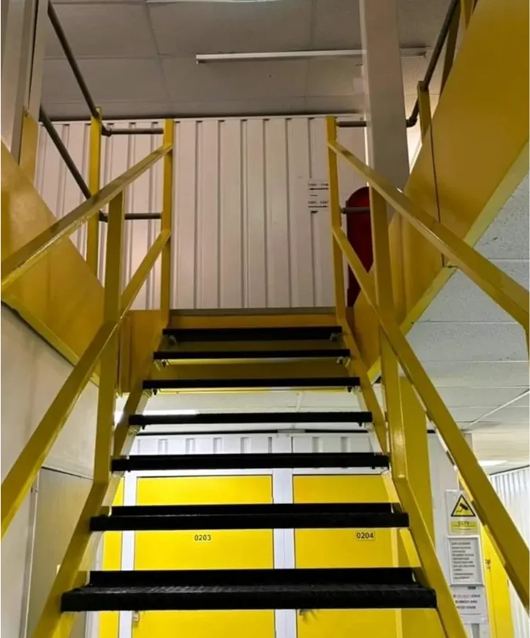 Indoor yellow metal staircase with black treads leading to an upper floor in an industrial or storage facility.