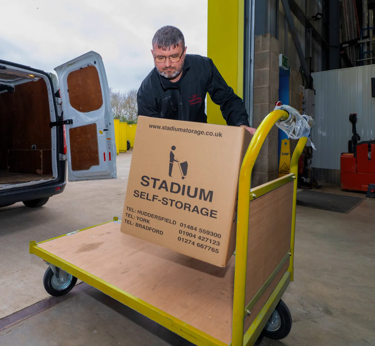 Man in black jacket loading a cardboard box labeled 'Stadium Self-Storage' onto a yellow trolley inside a storage facility.