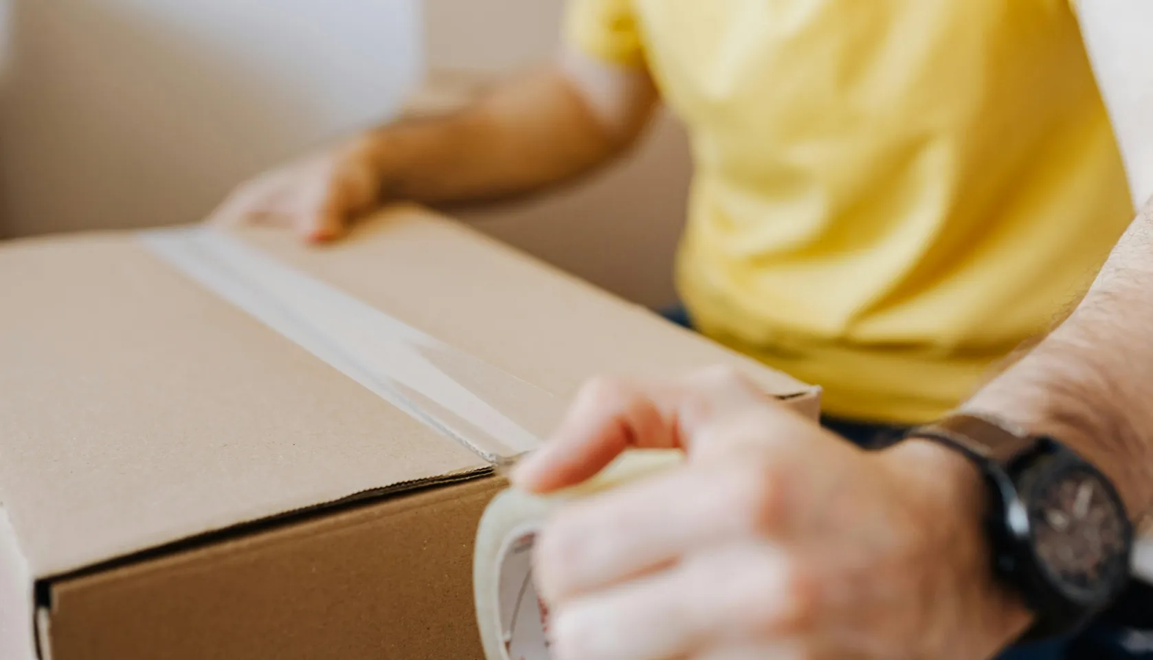 Person in a yellow shirt sealing a cardboard box with clear packing tape.