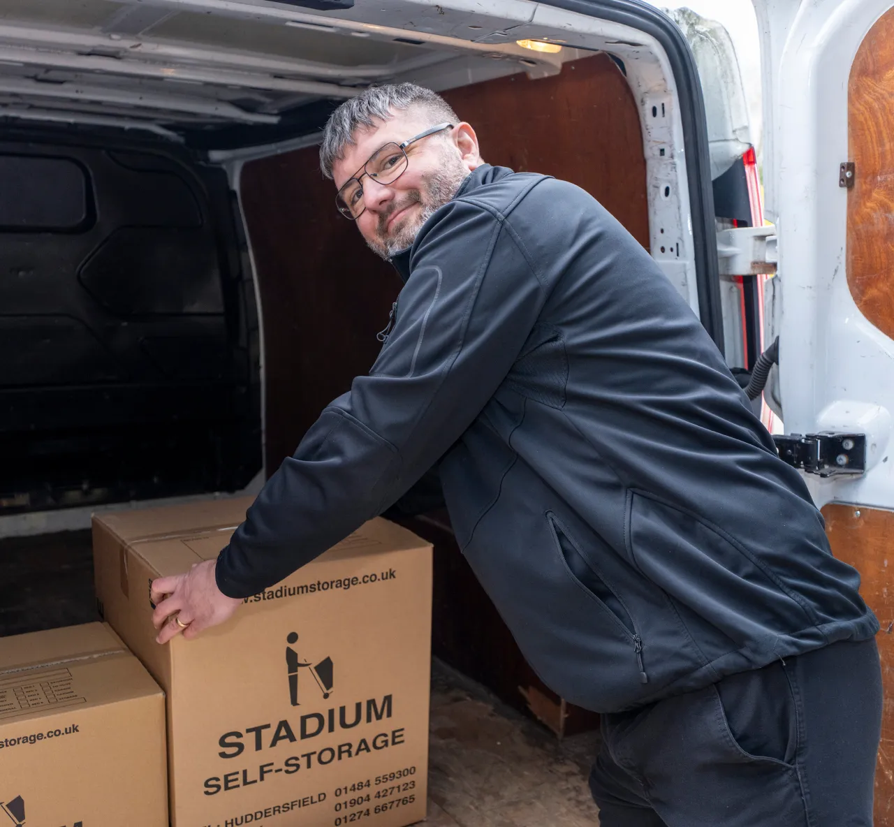 Man in black jacket loading or unloading cardboard boxes labeled Stadium Self-Storage from a white van.