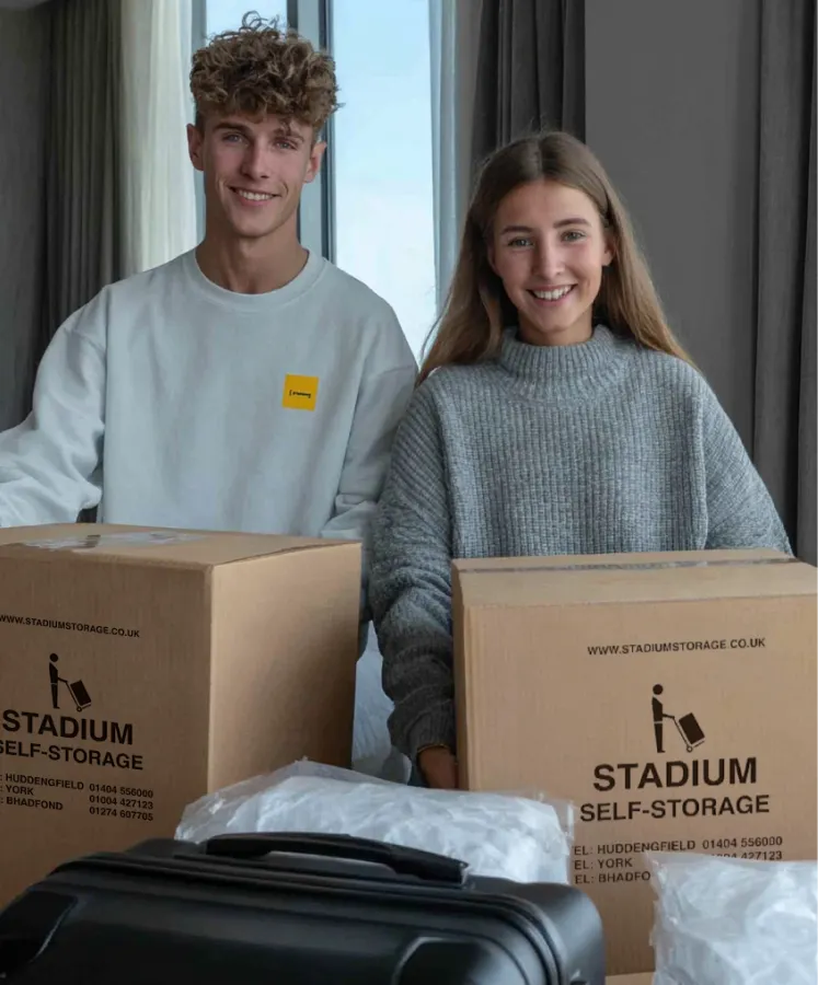 Smiling young man and woman sitting behind large cardboard storage boxes and luggage indoors.