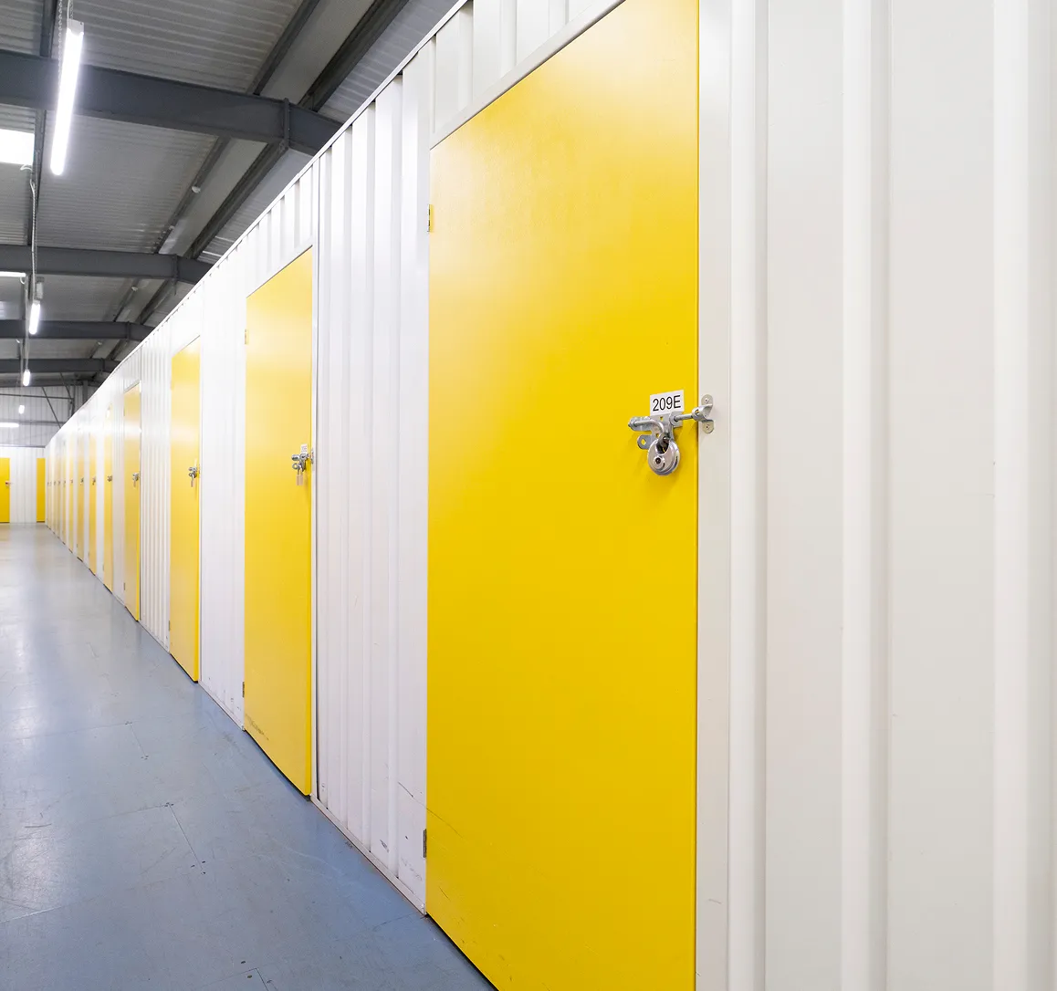 Indoor storage facility corridor with white walls and yellow metal storage unit doors, one labeled 209E with a padlock.