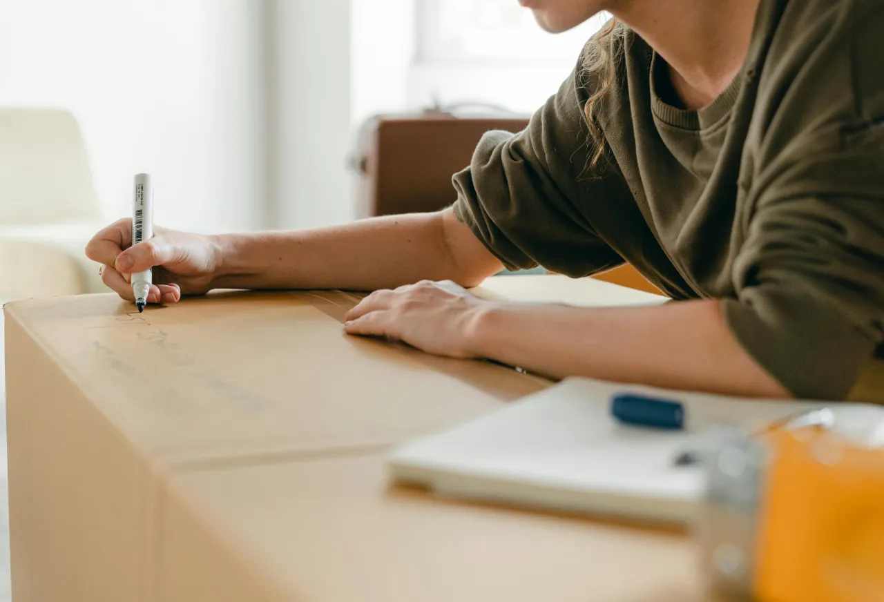 Person writing on a large cardboard box with a black marker while leaning on a table inside a room.