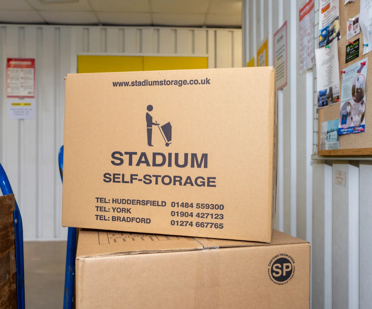 Two cardboard boxes stacked inside a storage unit, the top box with Stadium Self-Storage branding and contact details for Huddersfield, York, and Bradford.