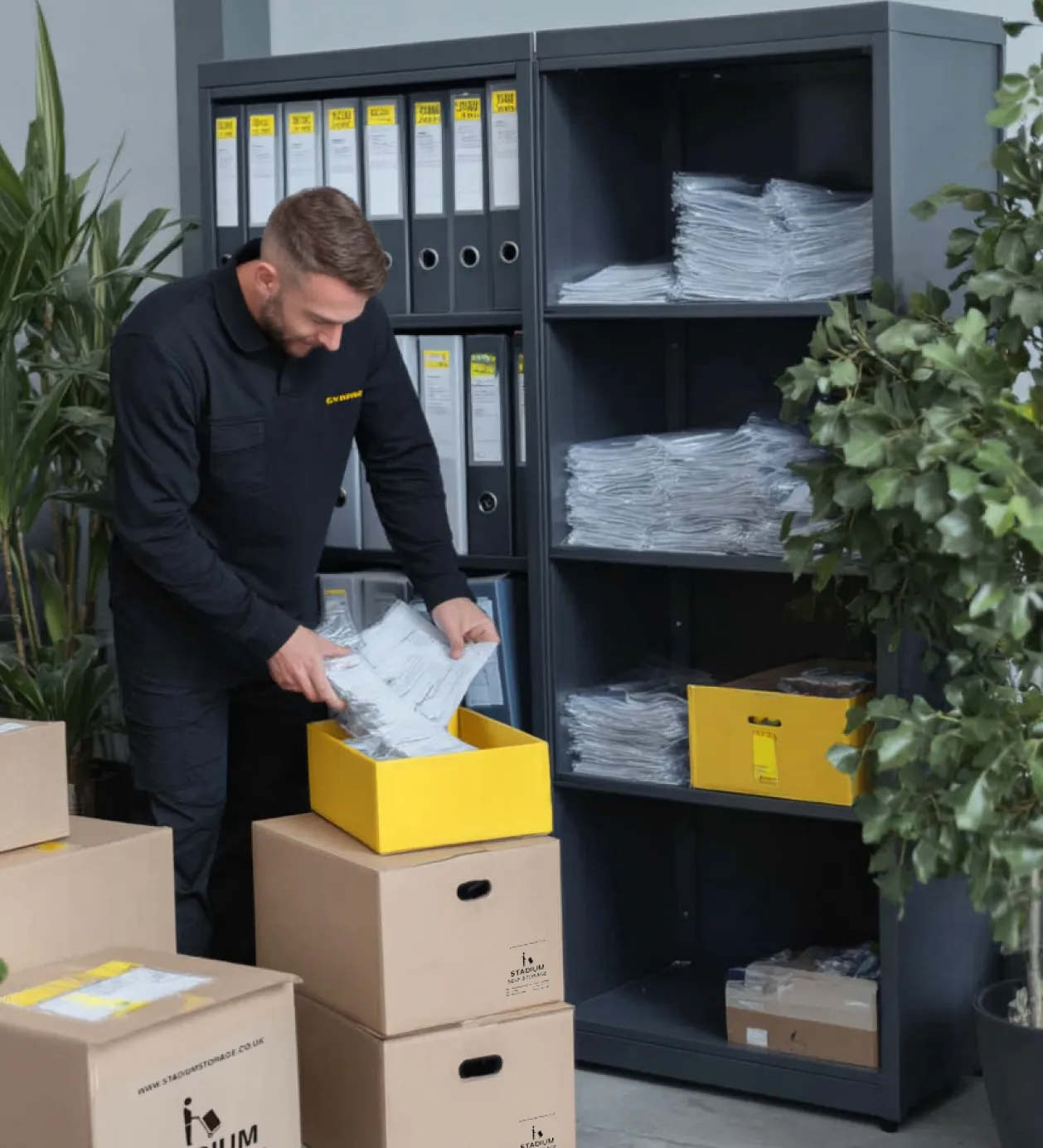 Man in black uniform organizing packaged documents into a yellow box surrounded by cardboard boxes and shelving with binders and stacks of paper.