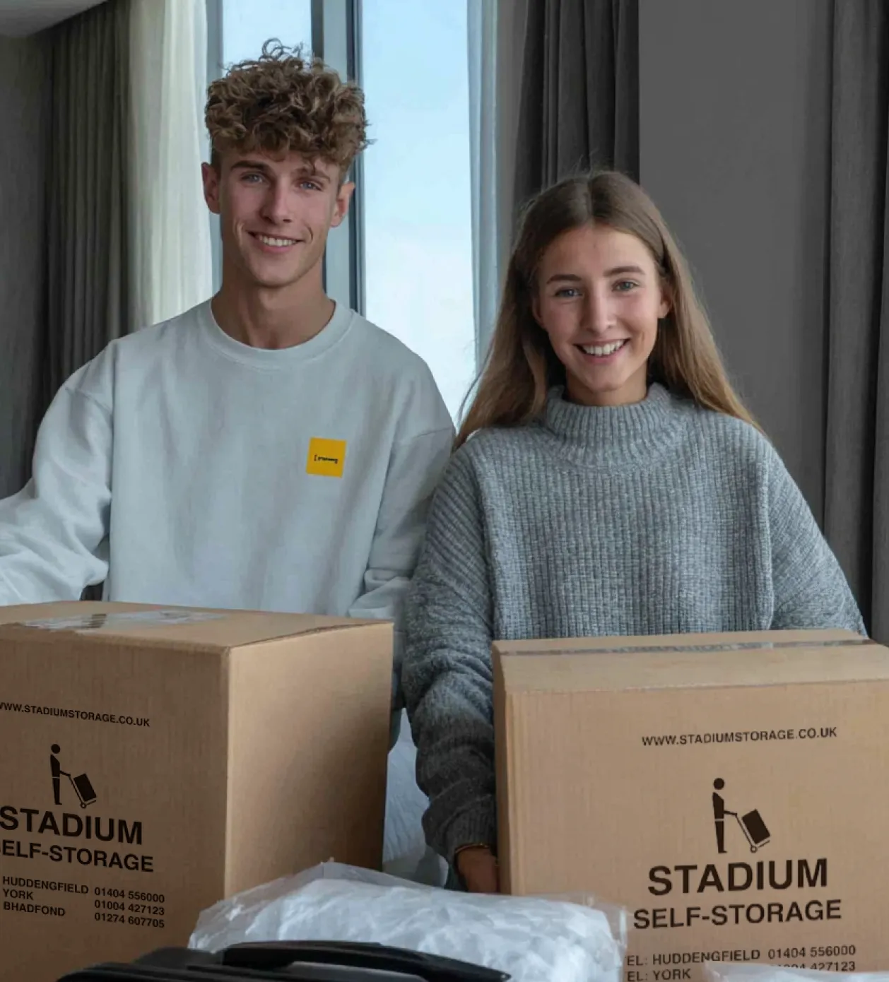 Young man and woman smiling indoors with two large cardboard boxes labeled Stadium Self-Storage.