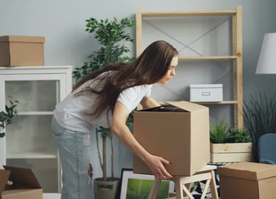 Woman with long brown hair lifting a large cardboard box indoors during moving or unpacking.