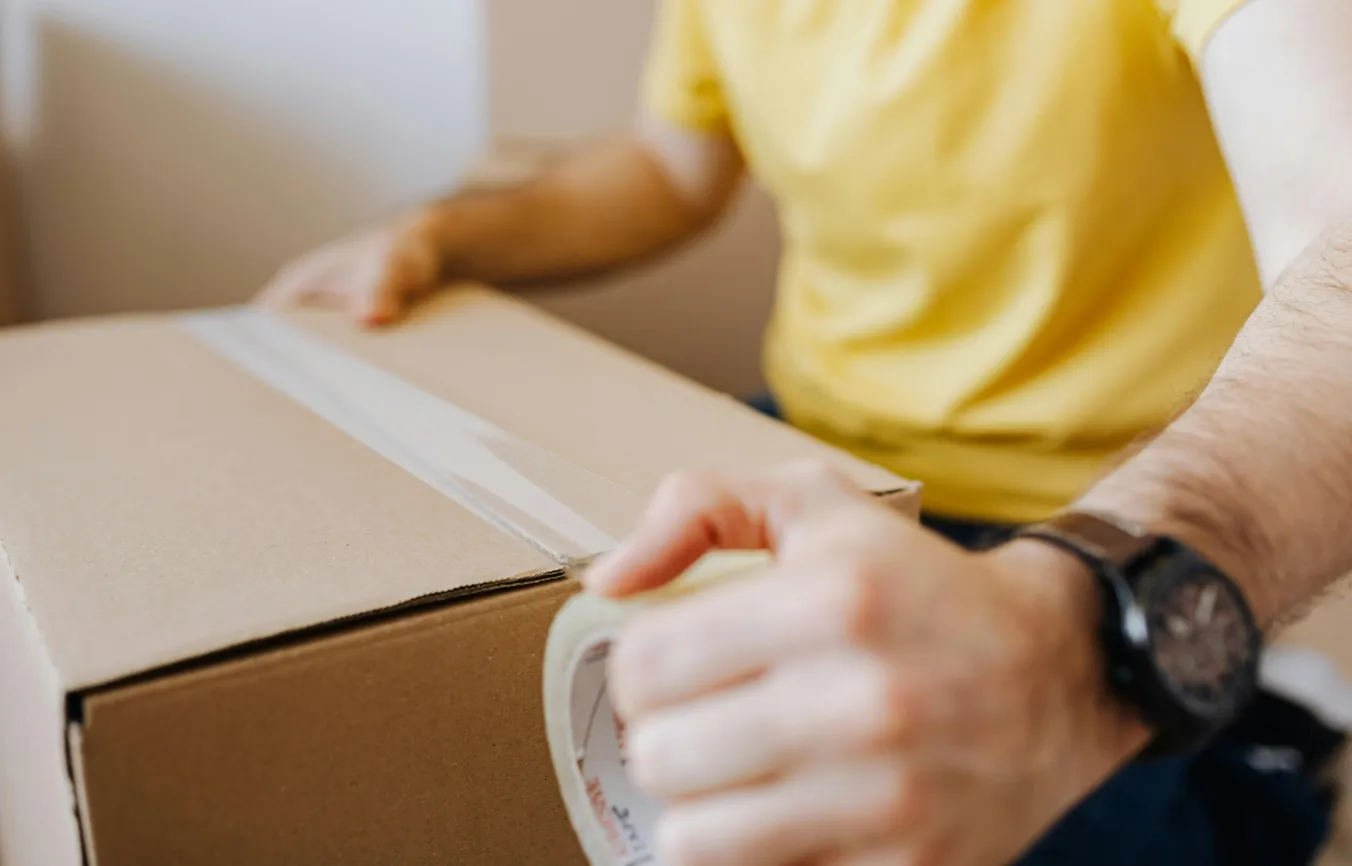 Person wearing a yellow shirt sealing a cardboard box with clear packing tape.