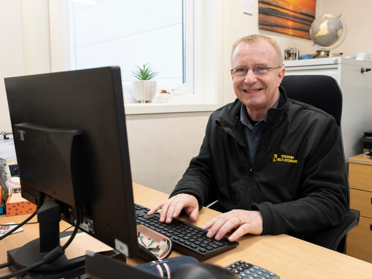Smiling man wearing a Stadium Self-Storage jacket types on a keyboard at an office desk.
