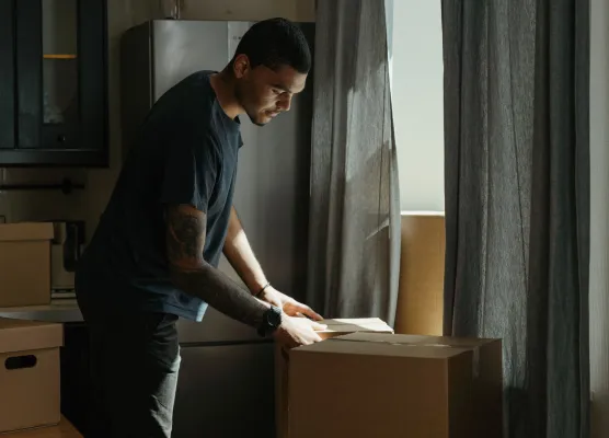 Man packing items into a cardboard box beside a window with gray curtains in a dim kitchen.