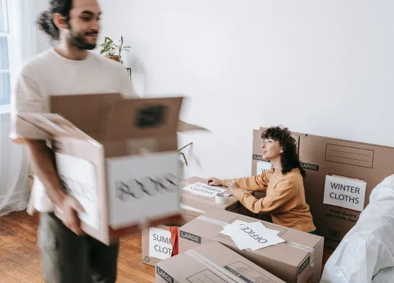 Woman sorting labeled moving boxes while man carries a box in a bright, white room.