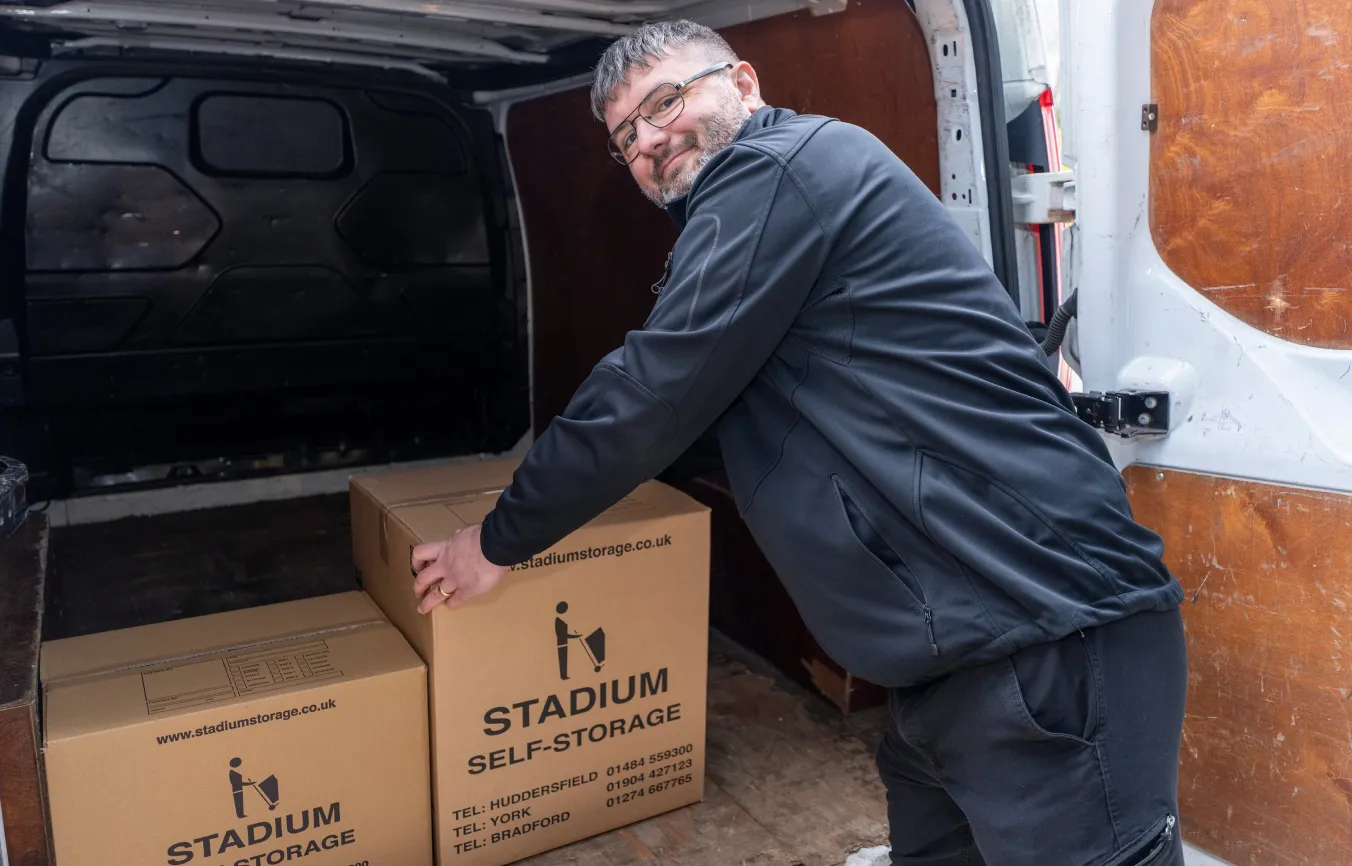 Man wearing glasses loading cardboard boxes labeled Stadium Self-Storage into the back of a van.