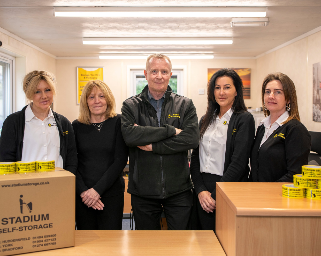 Five staff members in black Stadium Self-Storage uniforms standing behind a counter with branded packing tape.