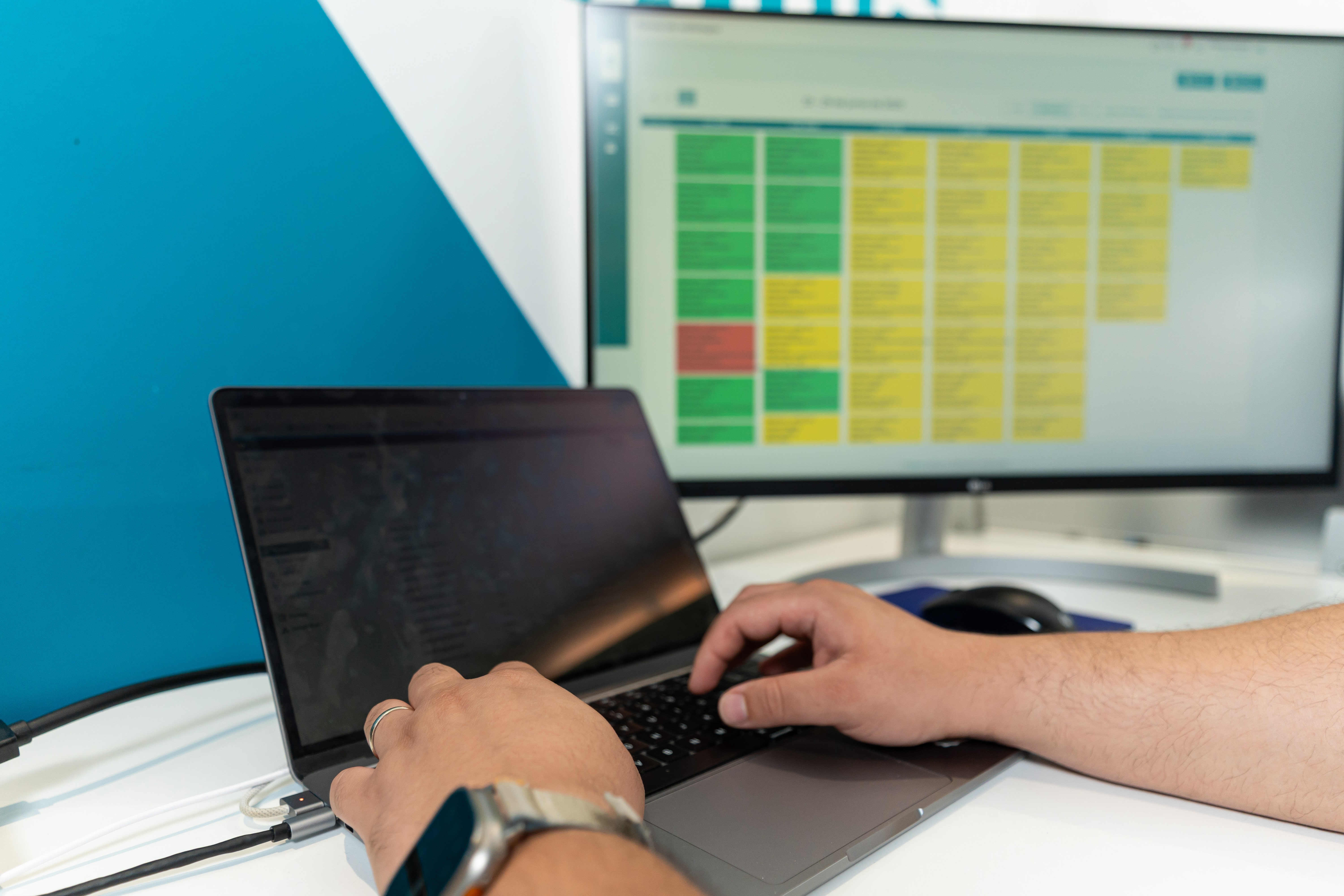 Persona tecleando en una computadora con un monitor al fondo que muestra un horario o una tabla codificada por colores.