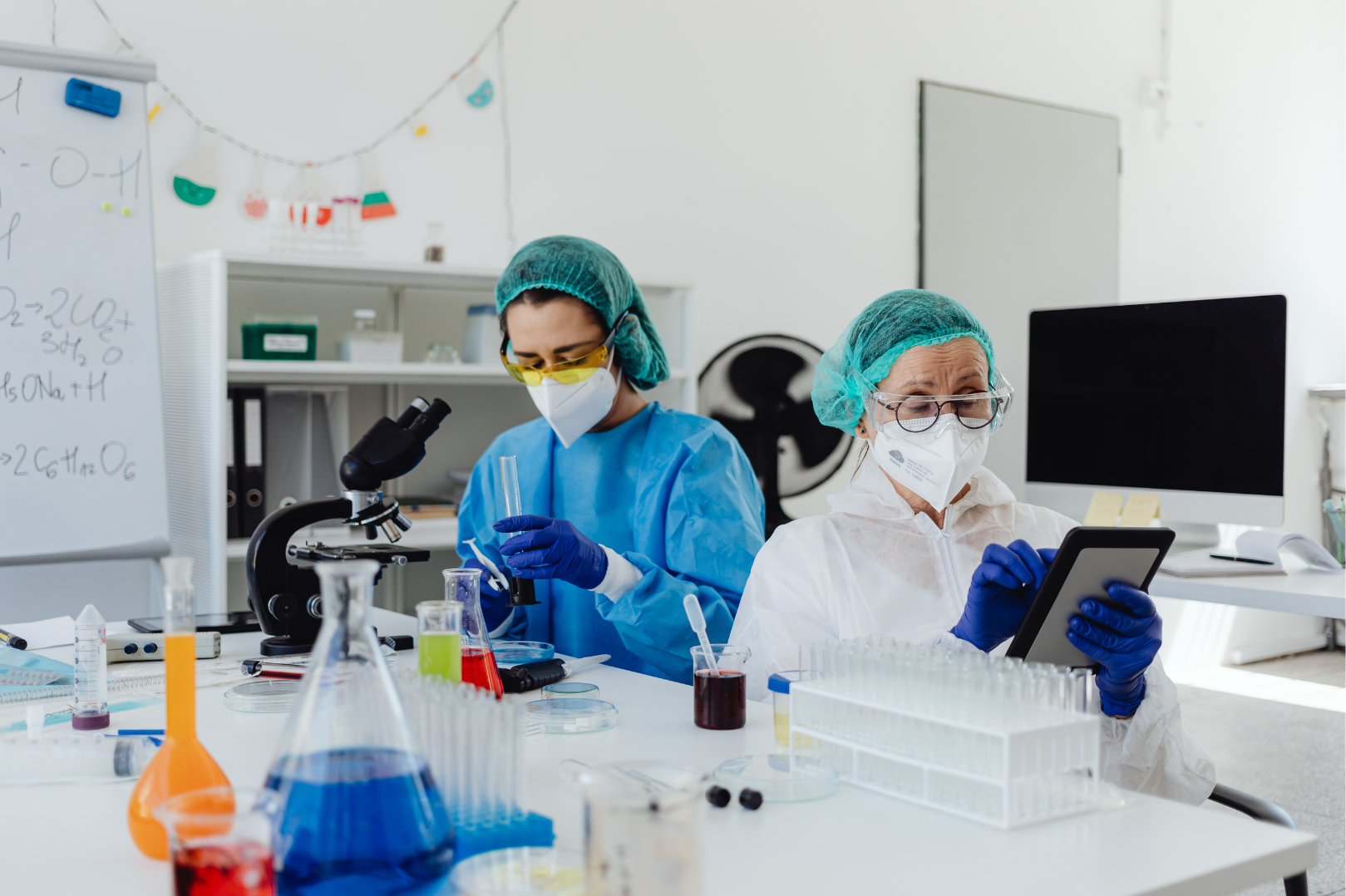 Dos científicos con equipo de protección trabajando en un laboratorio con tubos de ensayo, un microscopio y una tablet.