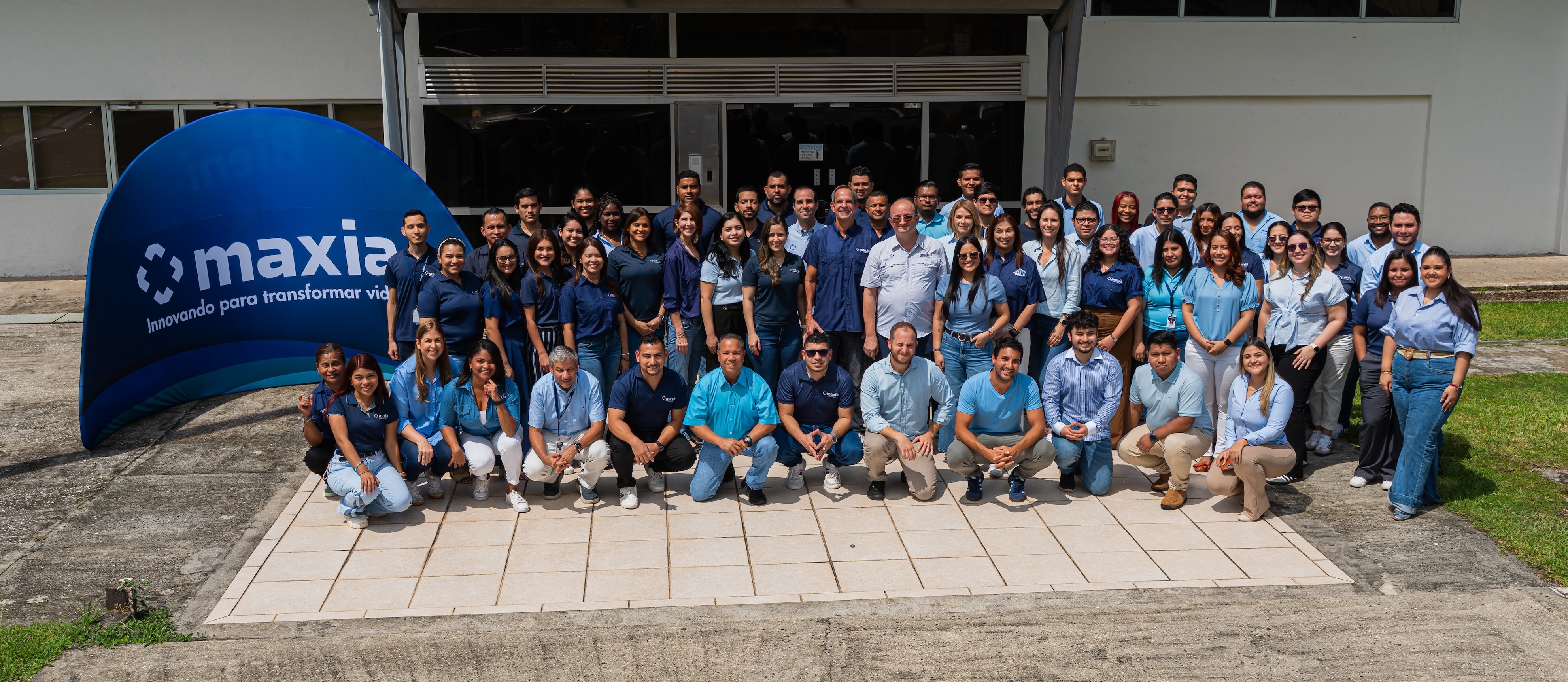 Un grupo numeroso de personas posando al aire libre frente a un edificio con una pancarta azul de Maxia en la que se lee «Innovando para transformar vidas».