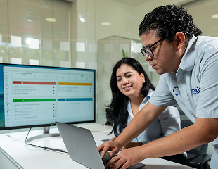 Dos compañeros de trabajo colaboran: uno escribe en una computadora portátil y el otro observa junto a un monitor en el que se muestra un panel de control de gestión de proyectos.