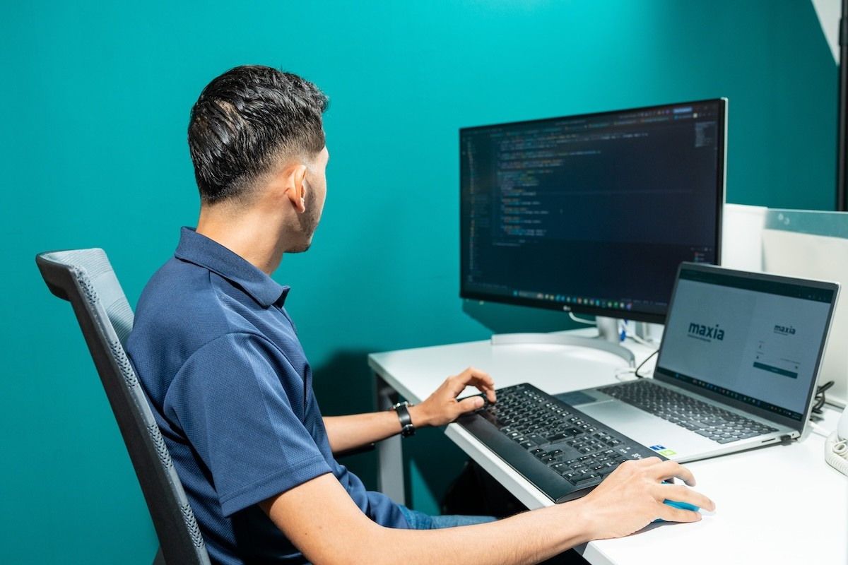Un hombre con una camisa azul sentado frente a un escritorio con monitores de computadora en los que se ven código y una pantalla de inicio de sesión.