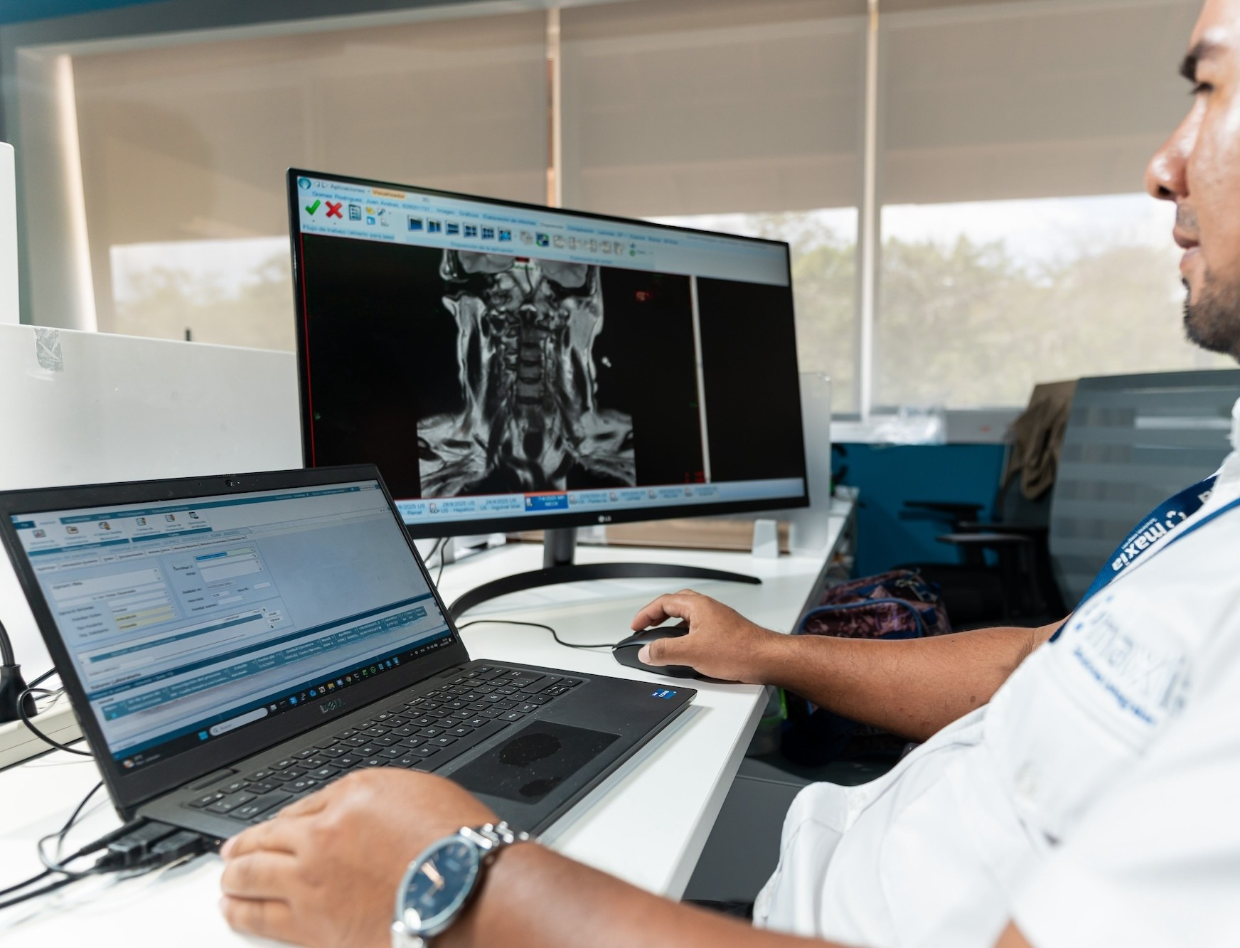 Persona utilizando una computadora y un mouse en un escritorio, con una imagen médica mostrada en un monitor al fondo.