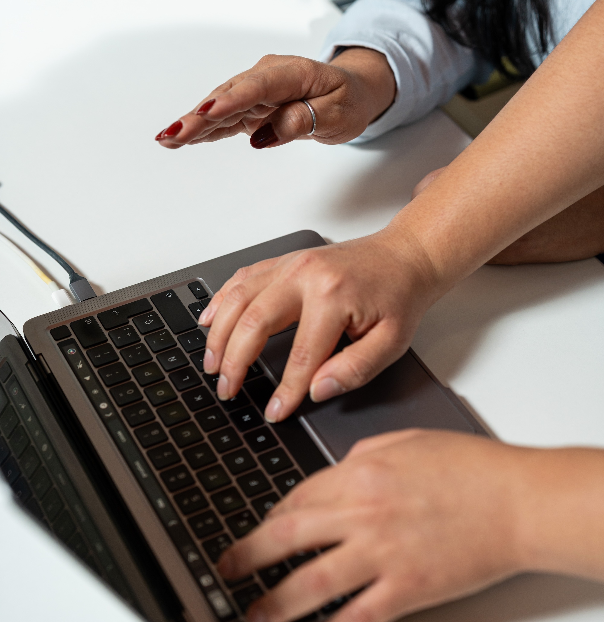 Dos personas trabajando juntas con las manos sobre el teclado de una computadora, sobre una mesa blanca.