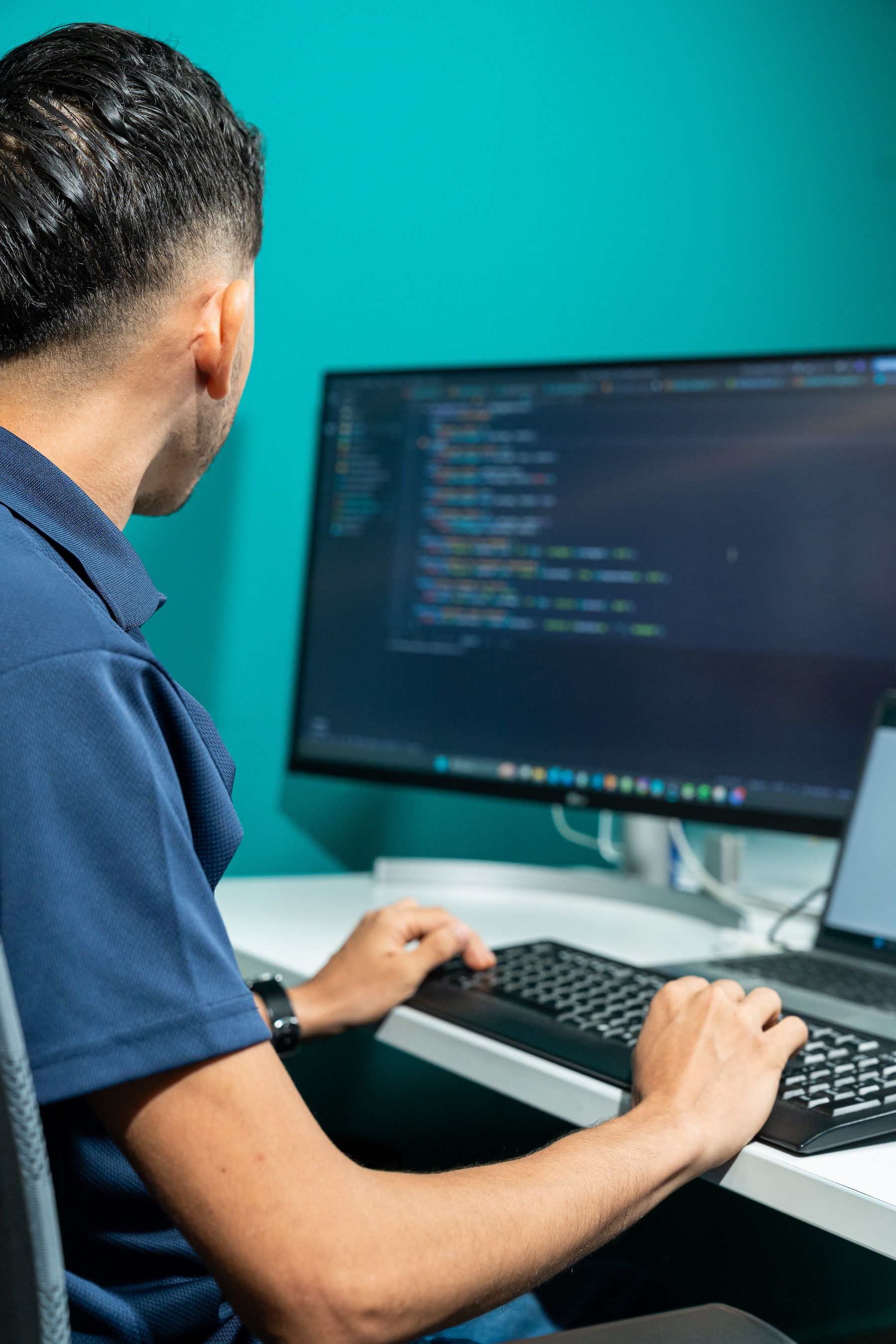 Un hombre con camisa azul trabajando en un código que se muestra en un monitor, sentado en un escritorio.