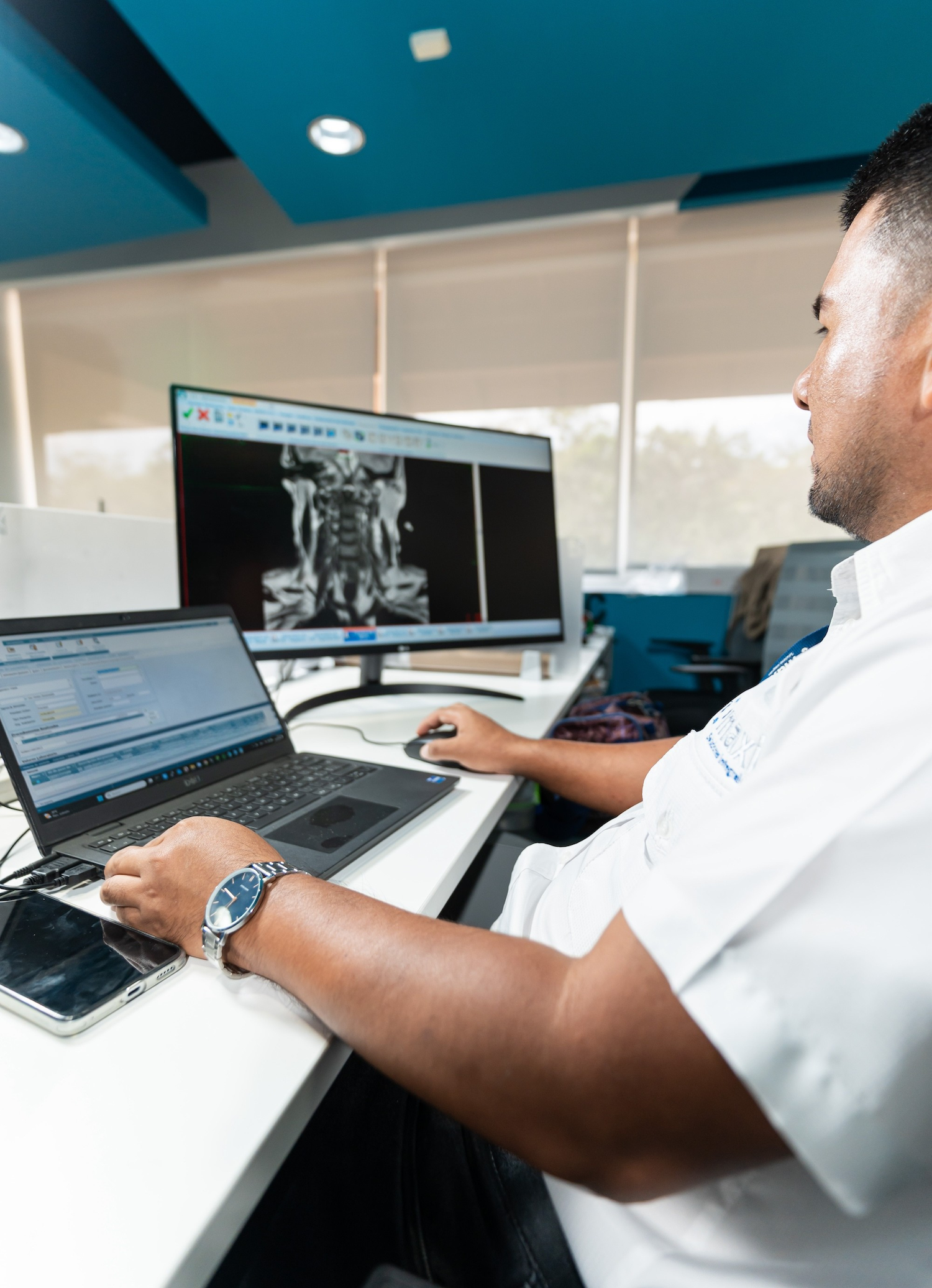 Un hombre con camisa blanca analiza imágenes médicas en el monitor de una computadora, mientras consulta los datos correspondientes en una computadora portátil, en una oficina.