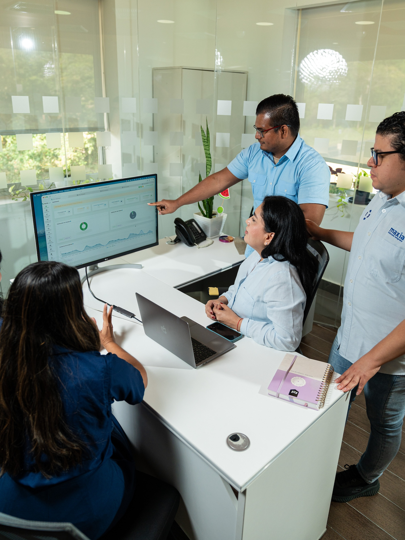 Cuatro compañeros de trabajo, en una oficina, comentan los datos que aparecen en la pantalla de una computadora, en la que se muestran tablas y gráficos.