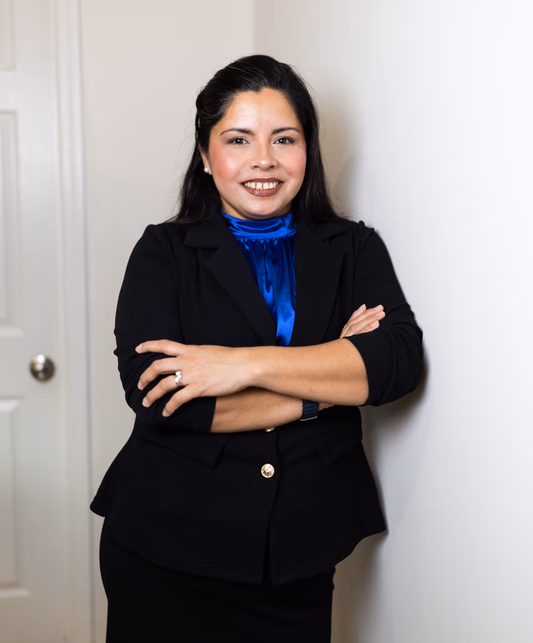 Smiling woman with dark hair crossed arms wearing a black blazer and blue blouse standing against a white wall.