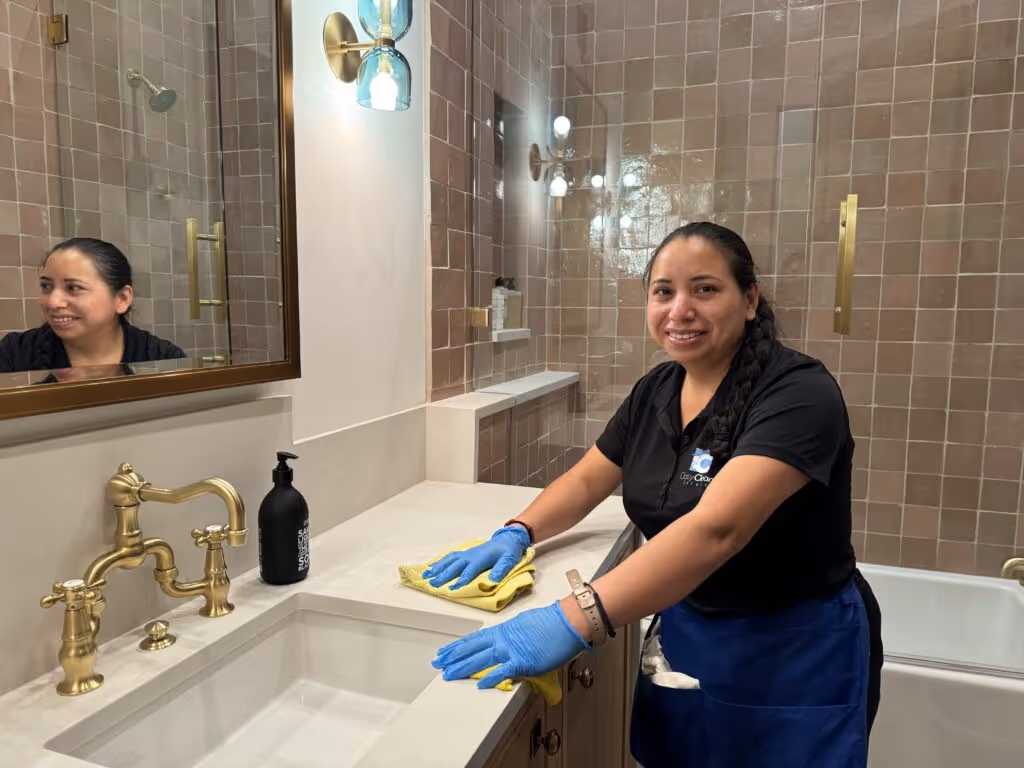 Woman in blue gloves and apron cleaning a bathroom sink countertop with a yellow cloth and smiling at the camera.