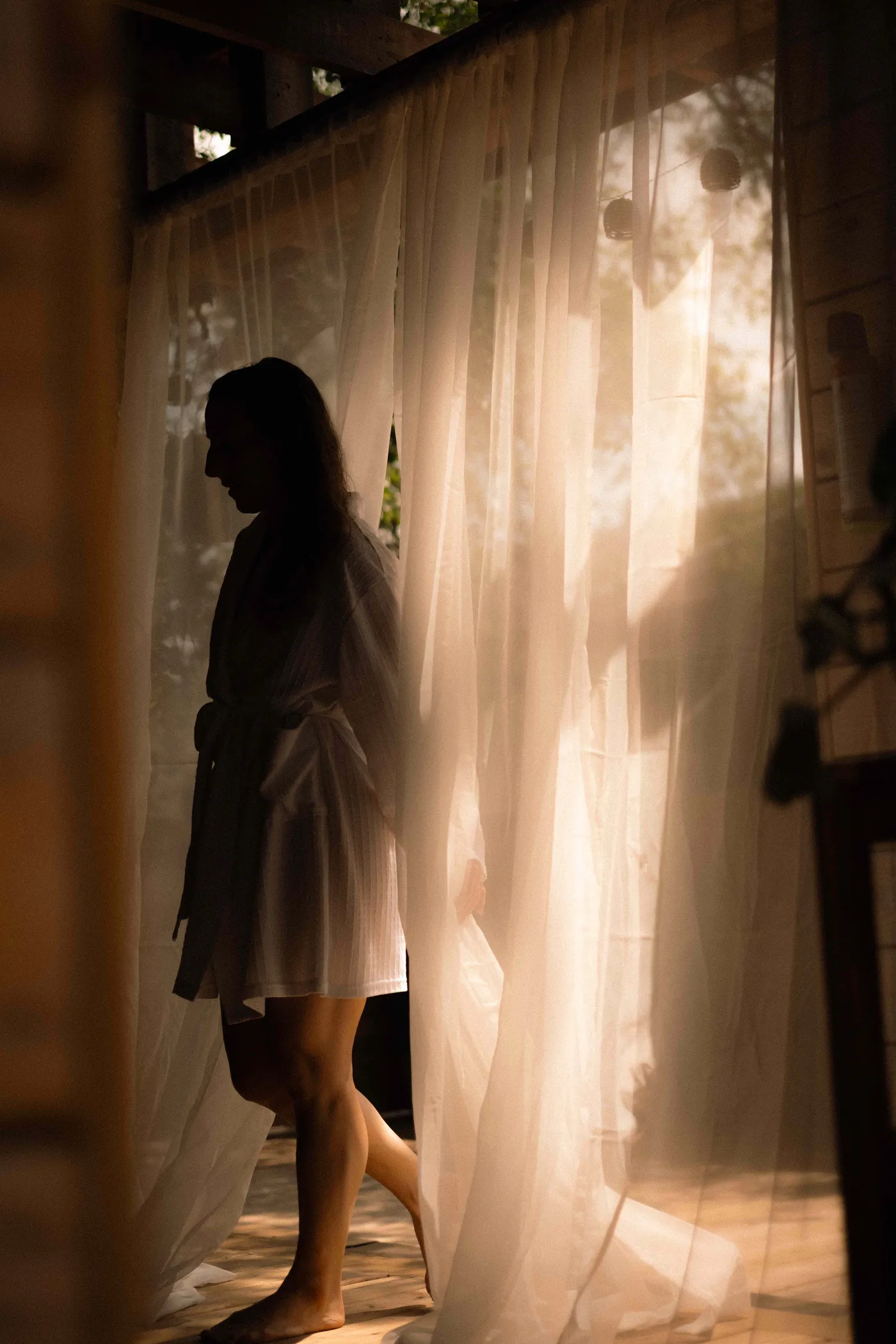 A guest in a robe walking past sunlit sheer curtains in a warm wood-lined room at Ralia