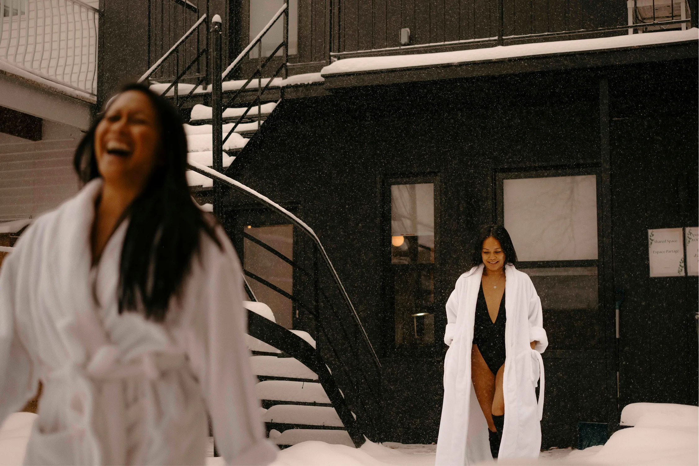 Two guests in white bathrobes laughing outside in falling snow during a winter stay with Ralia