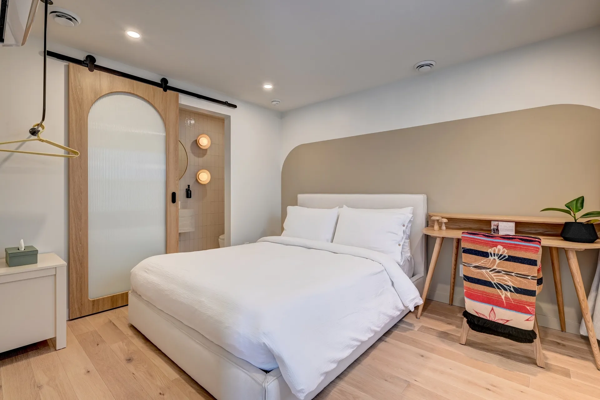 Sunlit guest room with a queen bed, wood desk and arched sliding door to the ensuite bathroom