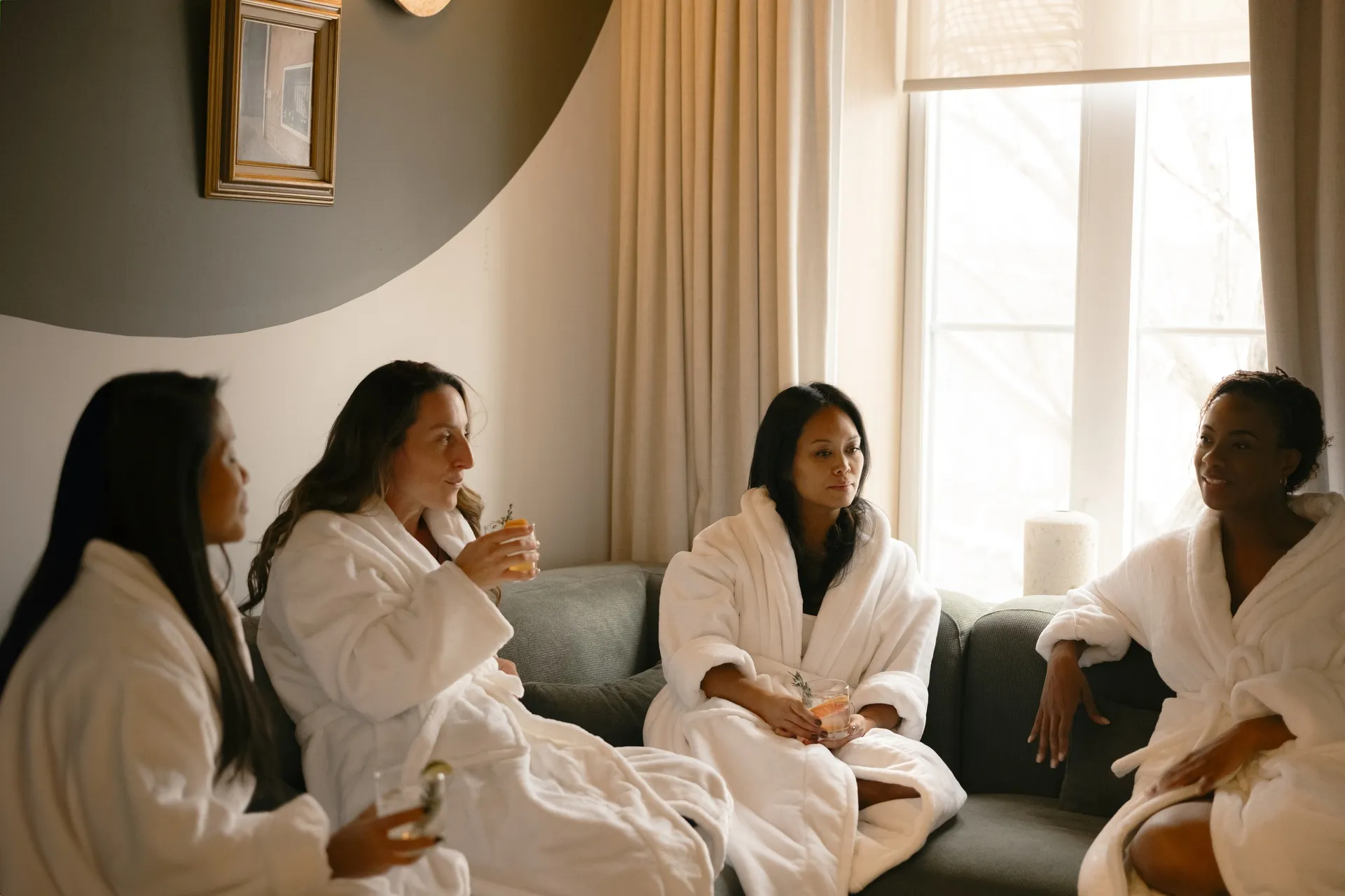 Four guests in white bathrobes relaxing and chatting on a sofa in a sunlit lounge at Ralia