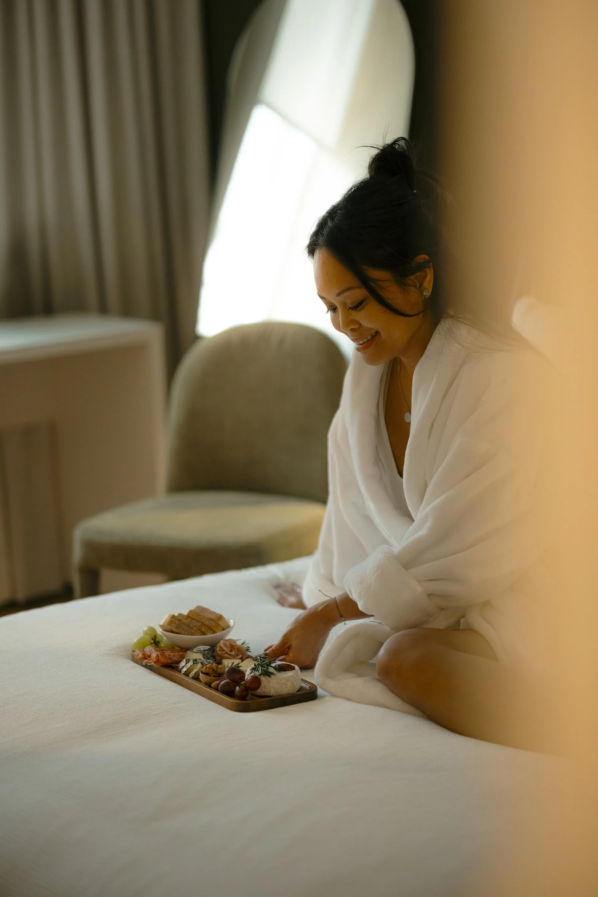 Woman in a white robe with a grazing board on the bed in a Ralia guest room