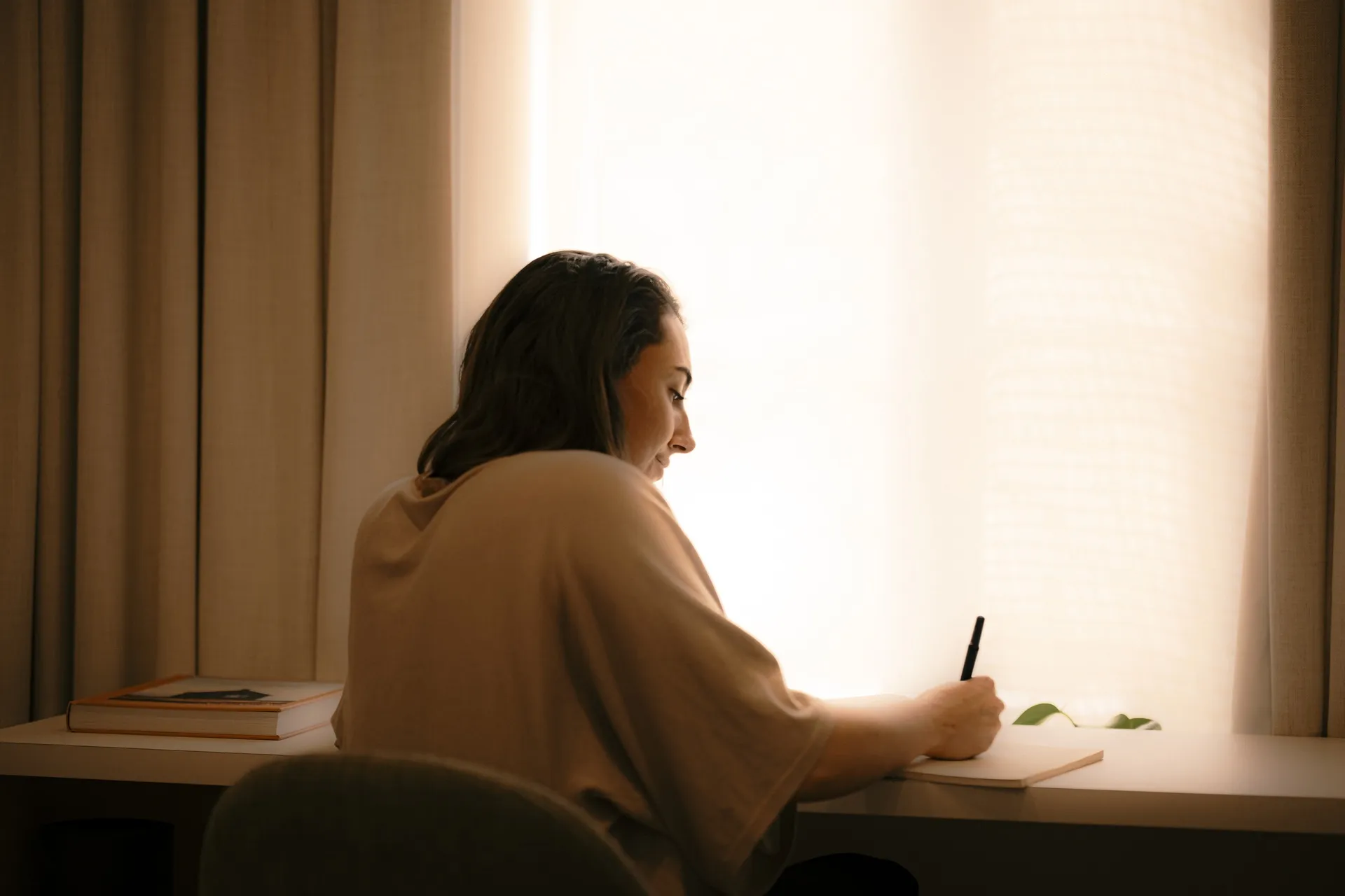 Woman journaling at a desk beside a softly lit curtained window during a quiet staycation