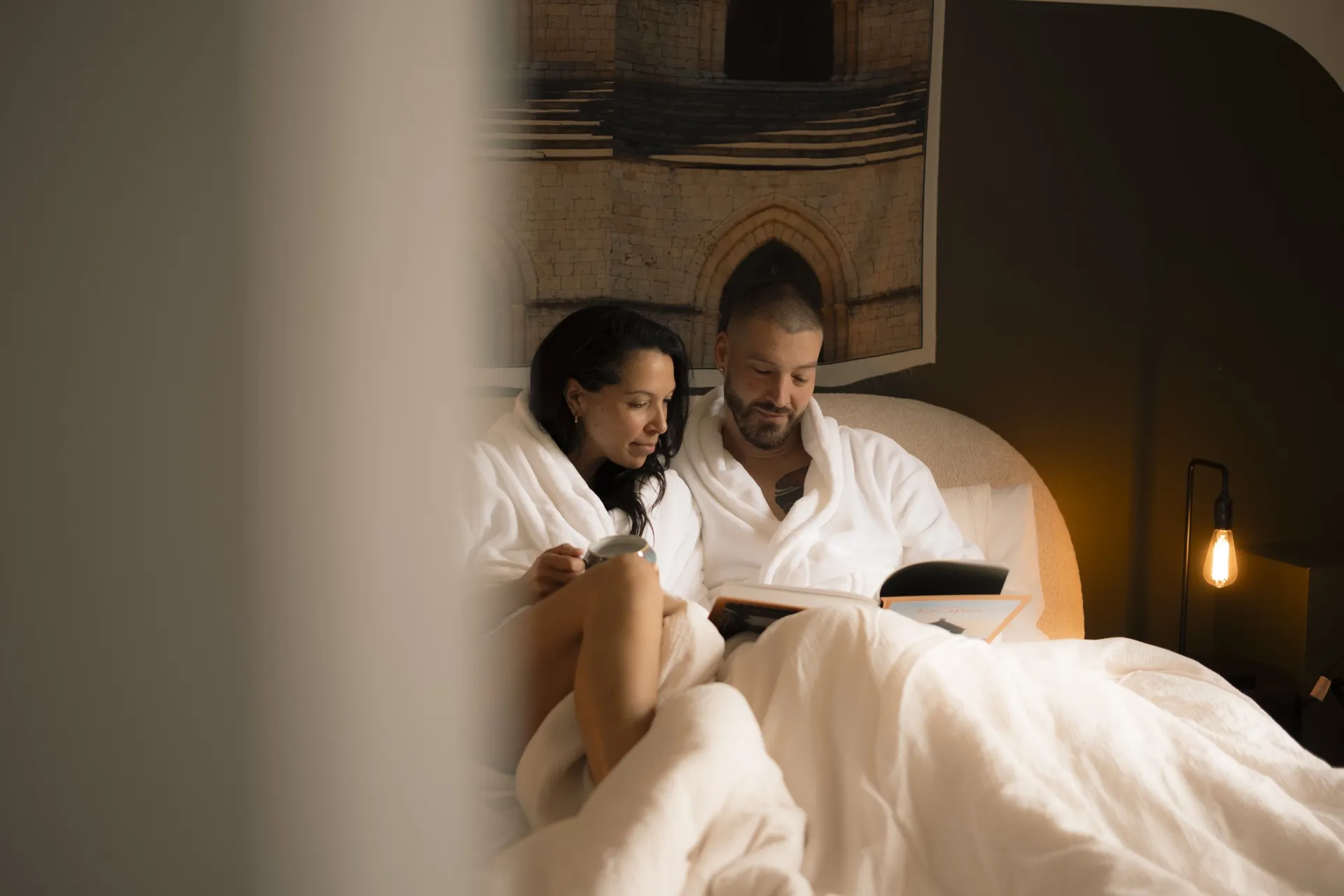 Couple in white bathrobes reading in bed in a warmly lit guest room at La Maison Ralia