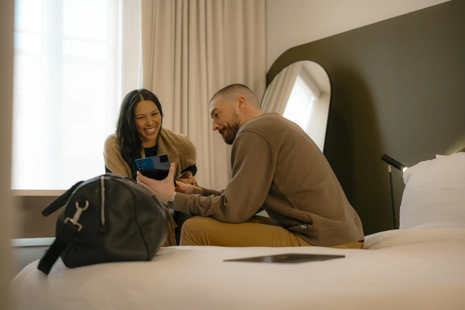 Two guests laughing together on the bed of a Ralia boutique hotel room, weekend bag beside them